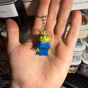 Könnte beinhalten: Ein Schlüsselanhänger mit einer Lego-artigen Alien-Figur. Der Alien hat einen grünen Kopf, drei Augen und einen blauen Körper mit einem rosa Kragen. Der Schlüsselanhänger ist an einem silbernen Metallring befestigt.