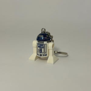 Könnte beinhalten: Ein weiß-blauer R2-D2-Schlüsselanhänger. Der Schlüsselanhänger zeigt eine Miniaturversion des ikonischen Droiden mit einer silbernen Kette und einem Schlüsselring. Der Droide hat eine blaue Kuppel und einen weißen Körper mit blauen und grauen Details.