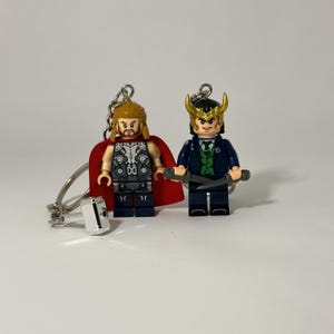 May include: Two Lego keychains featuring Thor and Loki figures. Thor has a red cape and a silver and black torso. Loki wears a dark blue suit with gold horns. Both figures have key rings attached.