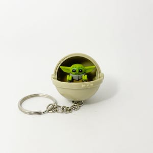 Könnte beinhalten: Ein Schlüsselanhänger mit einer grünen Alienfigur in einer beigefarbenen Kapsel. Der Außerirdische hat große Augen und spitze Ohren. Die Kapsel ist an einem silbernen Metall-Schlüsselring und einer Kette befestigt.