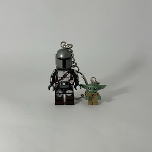 Könnte beinhalten: Ein Schlüsselanhänger mit zwei Lego-Figuren: Der Mandalorianer und Grogu. Die Mandalorianer-Figur hat einen silbernen Helm und eine detaillierte Rüstung. Grogu, auch bekannt als Baby Yoda, ist eine kleine, grüne Figur. Beide Figuren sind an einer silbernen Kette befestigt.