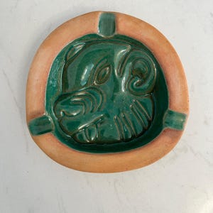 May include: Circular ceramic ashtray featuring a green ram's head design. The ashtray has a light brown rim and three green notches for cigarettes. A decorative item for the home.
