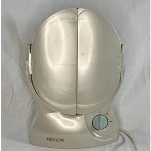 Puede incluir: Un espejo de tocador Revlon de color crema. El espejo tiene forma redondeada con dos paneles plegables. Un dial azul claro está en la base. El logotipo de Revlon está impreso en la base.