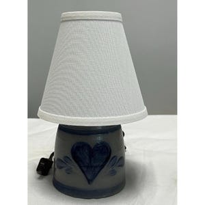 Pode incluir: Um candeeiro de mesa com base de cerâmica cinzenta decorada com um design de coração e folhas pintado em azul escuro. O candeeiro tem um abajur cónico de tecido branco e inclui um cabo elétrico preto com um pequeno interruptor na base.