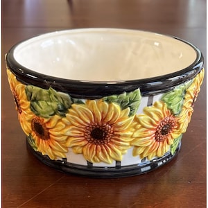 Peut inclure: Un bol en céramique avec un intérieur crème et un bord noir. L'extérieur présente un motif répété de tournesols jaunes avec des centres bruns et des feuilles vertes, sur un fond blanc avec des rayures verticales noires.