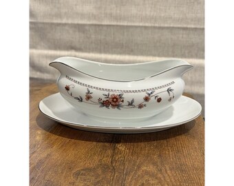 May include: White porcelain gravy boat with a matching saucer. The boat features a floral design in shades of pink, brown, and green, with a silver trim. The gravy boat is oval-shaped with a spout and a handle.