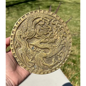 May include: A round, antique brass decorative plate featuring a detailed relief of a dragon and phoenix. The plate has an ornate border and is held in a person's hand against a background of green grass.