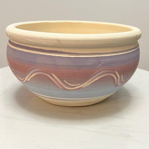 Puede incluir: Un cuenco de cerámica con borde e interior color crema. El exterior presenta bandas horizontales de color en tonos morados, rojos y azules, con un diseño ondulado color crema. El cuenco se utiliza probablemente con fines decorativos o como maceta.
