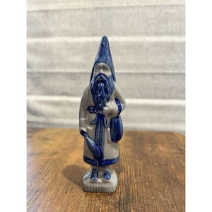 May include: A ceramic figurine of a bearded gnome in a blue pointed hat and robe, holding a bag and a bottle. The figurine is primarily white and blue, with intricate detailing. The gnome is standing on a small, rectangular base.