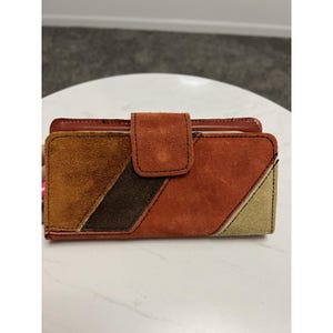 May include: A rectangular wallet with a suede exterior in shades of brown, tan, and beige. The wallet features a geometric design with a flap closure and a contrasting dark brown stripe. The wallet is made of suede and leather.