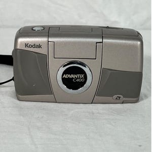 Puede incluir: Una cámara Kodak Advantix C400 plateada y gris. La cámara tiene forma rectangular con bordes redondeados y una lente en el centro. El logotipo de Kodak es visible en la esquina superior izquierda. Sobre una superficie blanca.