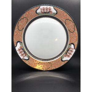 May include: A decorative ceramic plate with a white center and a patterned border. The border features a gold, orange, and black design with images of ships. The plate has a black rim and is likely for display or serving.