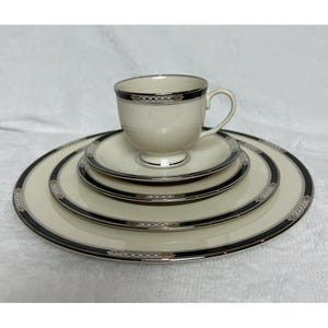 May include: Cream-colored teacup and saucer set with a black and silver border. The set includes a teacup, a saucer, a salad plate, and a dinner plate. The plates and cup have a matching design, creating a cohesive look.