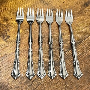 Può includere: Sei forchette d'argento con manici decorati sono disposte su una superficie di legno. Le forchette hanno tre rebbi e un motivo decorativo sui manici. I manici hanno una base svasata con un motivo a volute.
