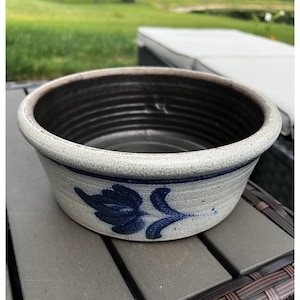 May include: A round ceramic bowl with a speckled light grey exterior and a dark brown interior. The exterior features a hand-painted dark blue floral motif and a thin horizontal blue line encircling the upper portion of the bowl's side.