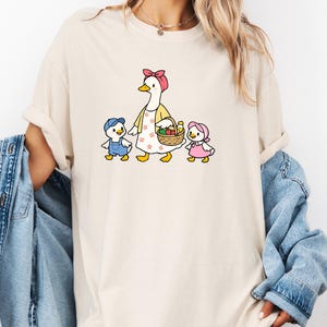 May include: A cream-coloured t-shirt with a cartoon goose wearing a pink bow and apron, leading two ducklings. The ducklings wear blue dungarees and a pink bonnet. The goose carries a basket of fruit.