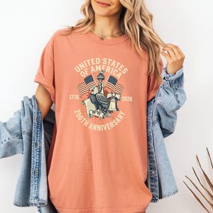 May include: A coral-colored t-shirt featuring a graphic commemorating the 250th anniversary of the United States of America. The design includes an eagle, flags, the Statue of Liberty, and the text "UNITED STATES OF AMERICA 250TH ANNIVERSARY".