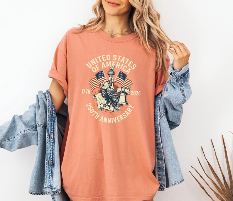 May include: A coral-colored t-shirt featuring a graphic commemorating the 250th anniversary of the United States of America. The design includes an eagle, flags, the Statue of Liberty, and the text "UNITED STATES OF AMERICA 250TH ANNIVERSARY".