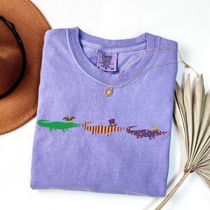 K&ouml;nnte beinhalten: Ein hellviolettes T-Shirt mit Rundhalsausschnitt und dem Text "COMFORT COLORS" auf dem Etikett. Das Shirt zeigt drei Cartoon-Alligatoren in Gr&uuml;n, Orange und Lila, die jeweils einen Hut tragen. Eine goldene Halskette liegt &uuml;ber dem Shirt.