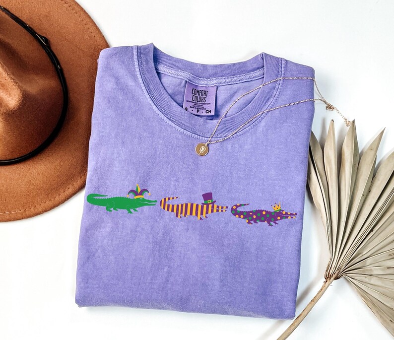 K&ouml;nnte beinhalten: Ein hellviolettes T-Shirt mit Rundhalsausschnitt und dem Text "COMFORT COLORS" auf dem Etikett. Das Shirt zeigt drei Cartoon-Alligatoren in Gr&uuml;n, Orange und Lila, die jeweils einen Hut tragen. Eine goldene Halskette liegt &uuml;ber dem Shirt.