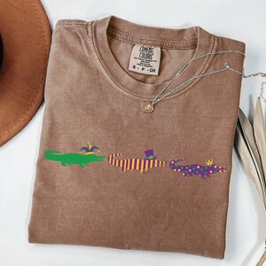 K&ouml;nnte beinhalten: Ein braunes Comfort Colors T-Shirt mit drei Cartoon-Alligatoren in Gr&uuml;n, Lila und Gelb. Die Alligatoren sind mit Accessoires im Mardi-Gras-Stil verziert. Eine silberne Halskette und ein brauner Hut sind ebenfalls sichtbar.