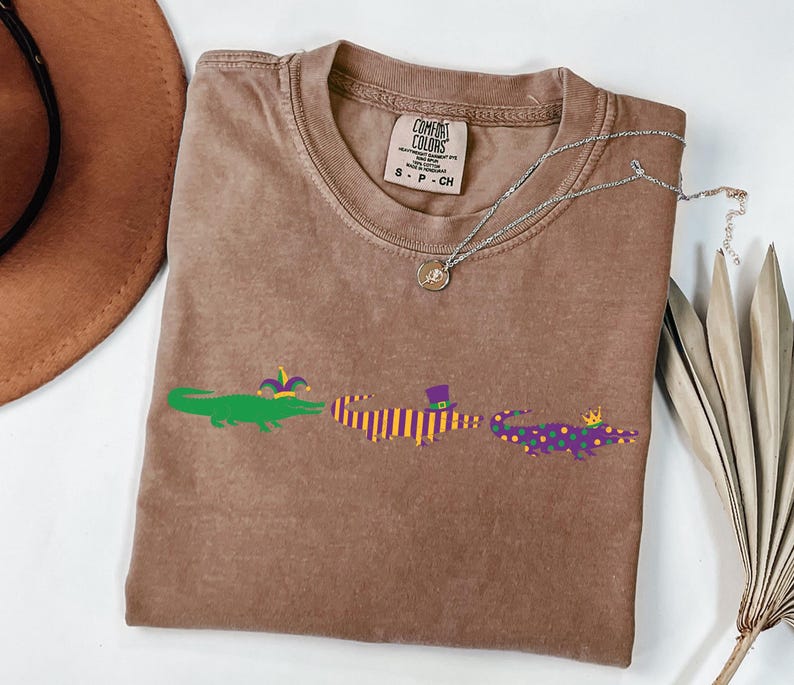 K&ouml;nnte beinhalten: Ein braunes Comfort Colors T-Shirt mit drei Cartoon-Alligatoren in Gr&uuml;n, Lila und Gelb. Die Alligatoren sind mit Accessoires im Mardi-Gras-Stil verziert. Eine silberne Halskette und ein brauner Hut sind ebenfalls sichtbar.