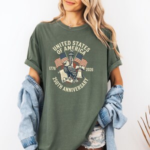 May include: Olive green t-shirt featuring a graphic commemorating the 250th anniversary of the United States of America. The design includes an eagle, American flags, and the text "United States of America 250th Anniversary 1776-2026."