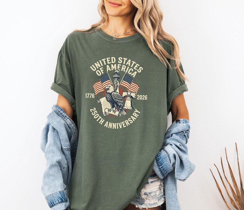 May include: Olive green t-shirt featuring a graphic commemorating the 250th anniversary of the United States of America. The design includes an eagle, American flags, and the text "United States of America 250th Anniversary 1776-2026."
