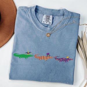 K&ouml;nnte beinhalten: Hellblaues T-Shirt mit drei Cartoon-Alligatoren in Gr&uuml;n, Lila und Gelb. Jeder Alligator tr&auml;gt einen festlichen Hut. Eine goldene Halskette und ein brauner Hut sind ebenfalls im Bild zu sehen.