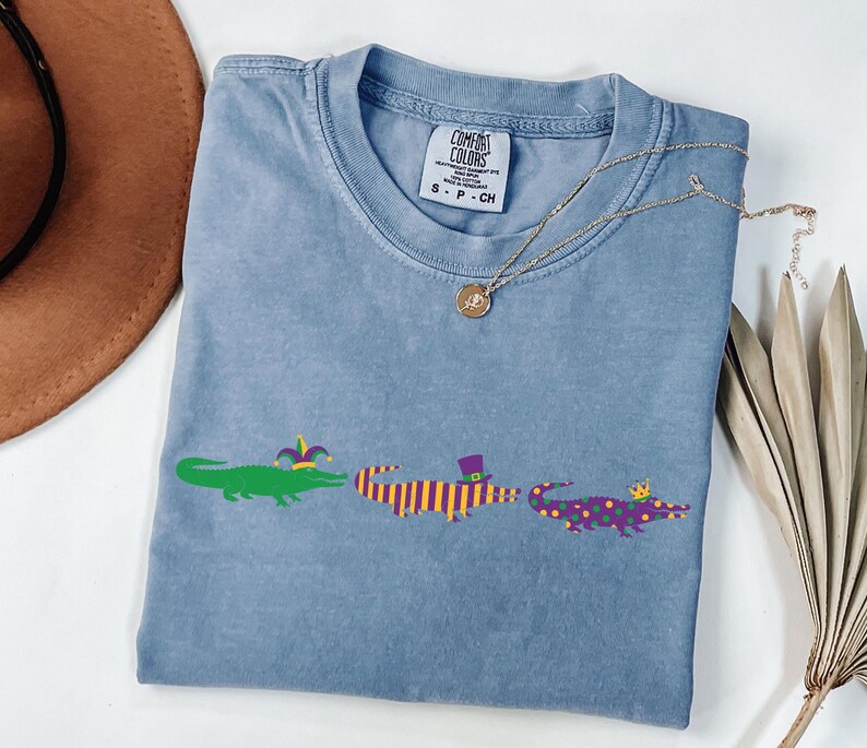 K&ouml;nnte beinhalten: Hellblaues T-Shirt mit drei Cartoon-Alligatoren in Gr&uuml;n, Lila und Gelb. Jeder Alligator tr&auml;gt einen festlichen Hut. Eine goldene Halskette und ein brauner Hut sind ebenfalls im Bild zu sehen.