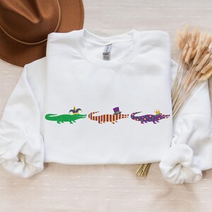 K&ouml;nnte beinhalten: Wei&szlig;es Sweatshirt mit drei Cartoon-Alligatoren in einer Reihe, die jeweils mit Mardi Gras-Dekorationen geschm&uuml;ckt sind. Die Alligatoren sind gr&uuml;n, orange und lila, mit H&uuml;ten und Kronen.