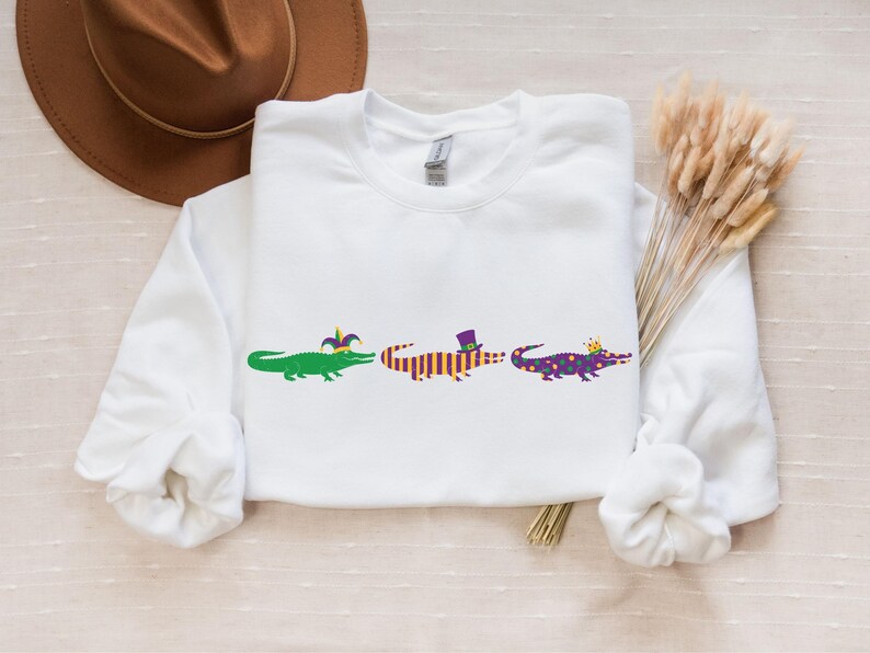 K&ouml;nnte beinhalten: Wei&szlig;es Sweatshirt mit drei Cartoon-Alligatoren in einer Reihe, die jeweils mit Mardi Gras-Dekorationen geschm&uuml;ckt sind. Die Alligatoren sind gr&uuml;n, orange und lila, mit H&uuml;ten und Kronen.