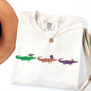 K&ouml;nnte beinhalten: Cremefarbenes T-Shirt mit drei Cartoon-Alligatoren in einer Reihe. Die Alligatoren sind gr&uuml;n, lila und gelb und tragen festliche H&uuml;te und Kronen. Das Shirt hat das Markenetikett Comfort Colors.