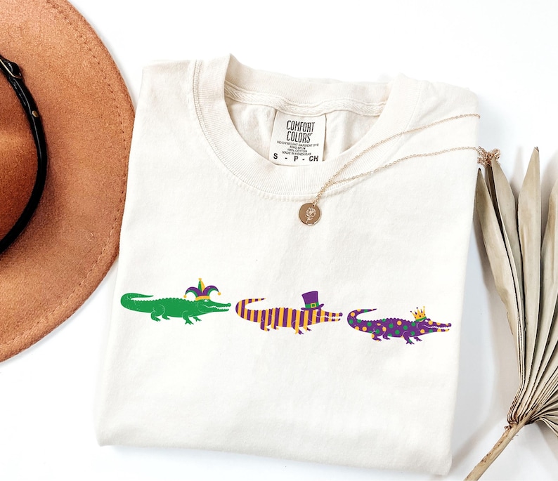 K&ouml;nnte beinhalten: Cremefarbenes T-Shirt mit drei Cartoon-Alligatoren in einer Reihe. Die Alligatoren sind gr&uuml;n, lila und gelb und tragen festliche H&uuml;te und Kronen. Das Shirt hat das Markenetikett Comfort Colors.