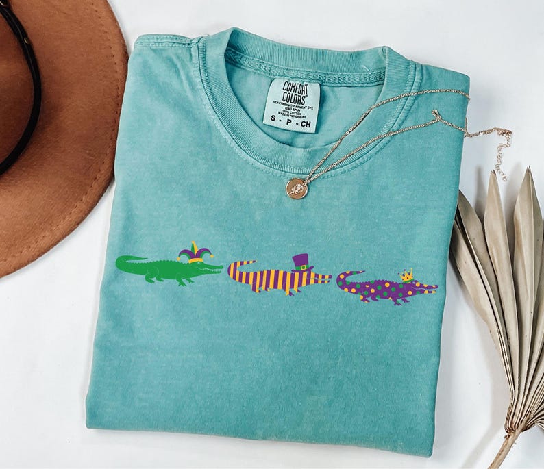 K&ouml;nnte beinhalten: Ein t&uuml;rkisfarbenes Comfort Colors T-Shirt mit drei Cartoon-Alligatoren mit H&uuml;ten. Die Alligatoren sind gr&uuml;n, lila und gelb, mit lila und gelben Streifen und gepunkteten Mustern. Eine goldene Halskette und ein brauner Hut sind im Hintergrund.
