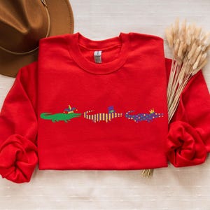 K&ouml;nnte beinhalten: Ein rotes Sweatshirt mit drei Cartoon-Alligatoren, die H&uuml;te tragen. Die Alligatoren sind gr&uuml;n, gelb und lila. Das Sweatshirt wird mit einem braunen Hut und Trockenblumen pr&auml;sentiert.