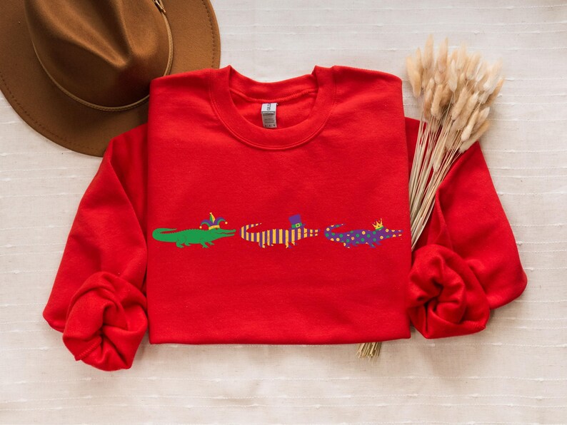 K&ouml;nnte beinhalten: Ein rotes Sweatshirt mit drei Cartoon-Alligatoren, die H&uuml;te tragen. Die Alligatoren sind gr&uuml;n, gelb und lila. Das Sweatshirt wird mit einem braunen Hut und Trockenblumen pr&auml;sentiert.