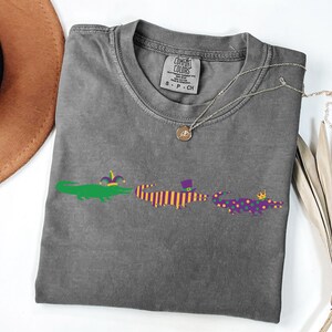 K&ouml;nnte beinhalten: Ein graues T-Shirt mit drei Cartoon-Alligatoren in Gr&uuml;n, Lila und Orange, die jeweils einen festlichen Hut tragen. Eine silberne Halskette mit Anh&auml;nger liegt auf dem Shirt. Das Shirt ist auf einer wei&szlig;en Oberfl&auml;che gefaltet.