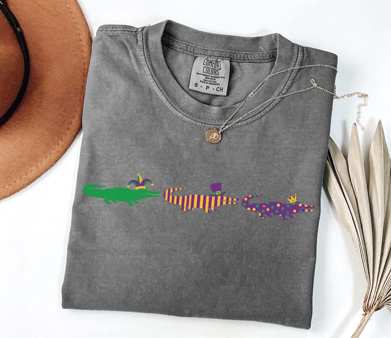 K&ouml;nnte beinhalten: Ein graues T-Shirt mit drei Cartoon-Alligatoren in Gr&uuml;n, Lila und Orange, die jeweils einen festlichen Hut tragen. Eine silberne Halskette mit Anh&auml;nger liegt auf dem Shirt. Das Shirt ist auf einer wei&szlig;en Oberfl&auml;che gefaltet.