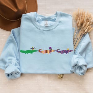 K&ouml;nnte beinhalten: Hellblaues Sweatshirt mit drei Cartoon-Alligatoren in einer Reihe. Jeder Alligator ist mit Mardi-Gras-Accessoires wie H&uuml;ten und festlichen Farben geschm&uuml;ckt. Ein brauner Hut und Trockenblumen befinden sich im Hintergrund.
