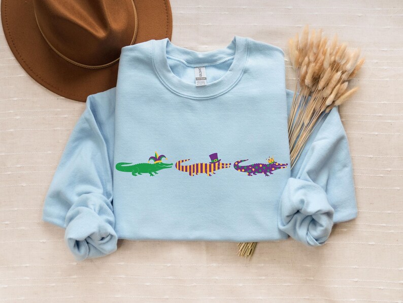 K&ouml;nnte beinhalten: Hellblaues Sweatshirt mit drei Cartoon-Alligatoren in einer Reihe. Jeder Alligator ist mit Mardi-Gras-Accessoires wie H&uuml;ten und festlichen Farben geschm&uuml;ckt. Ein brauner Hut und Trockenblumen befinden sich im Hintergrund.