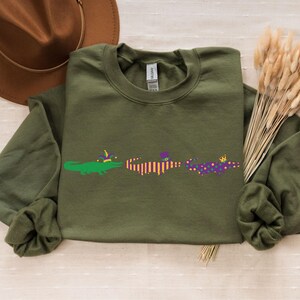 K&ouml;nnte beinhalten: Olivgr&uuml;nes Sweatshirt mit drei Cartoon-Alligatoren, die mit Mardi-Gras-Dekorationen verziert sind. Ein brauner Hut und Trockenblumen sind im Hintergrund. Ein l&auml;ssiges Kleidungsst&uuml;ck.
