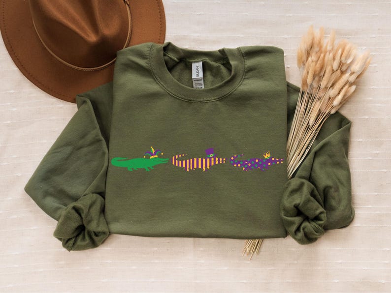 K&ouml;nnte beinhalten: Olivgr&uuml;nes Sweatshirt mit drei Cartoon-Alligatoren, die mit Mardi-Gras-Dekorationen verziert sind. Ein brauner Hut und Trockenblumen sind im Hintergrund. Ein l&auml;ssiges Kleidungsst&uuml;ck.