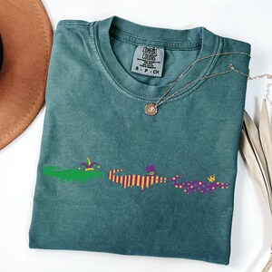 K&ouml;nnte beinhalten: T&uuml;rkisfarbenes Comfort Colors T-Shirt mit Rundhalsausschnitt und kurzen &Auml;rmeln. Das Shirt zeigt eine Grafik von drei Alligatoren in Mardi Gras-Kleidung. Eine goldene Halskette mit Anh&auml;nger ist auf dem Shirt drapiert.