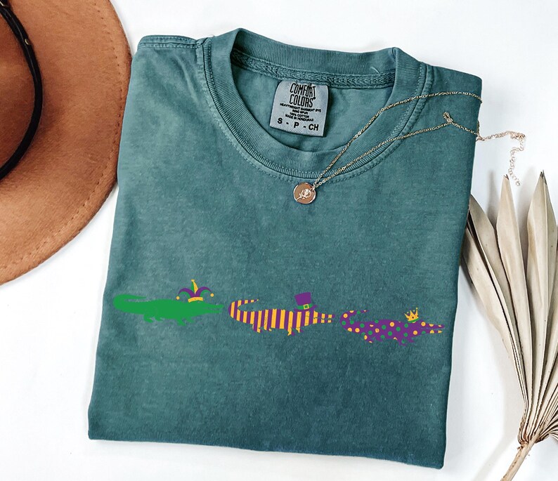 K&ouml;nnte beinhalten: T&uuml;rkisfarbenes Comfort Colors T-Shirt mit Rundhalsausschnitt und kurzen &Auml;rmeln. Das Shirt zeigt eine Grafik von drei Alligatoren in Mardi Gras-Kleidung. Eine goldene Halskette mit Anh&auml;nger ist auf dem Shirt drapiert.