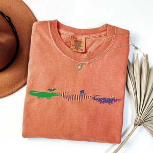 K&ouml;nnte beinhalten: Ein korallenfarbenes Comfort Colors T-Shirt mit drei Cartoon-Alligatoren in Gr&uuml;n, Lila und Lila mit goldenen Kronen. Die Alligatoren sind mit Accessoires zum Thema Mardi Gras verziert. Eine goldene Halskette und ein brauner Hut sind ebenfalls im Bild.