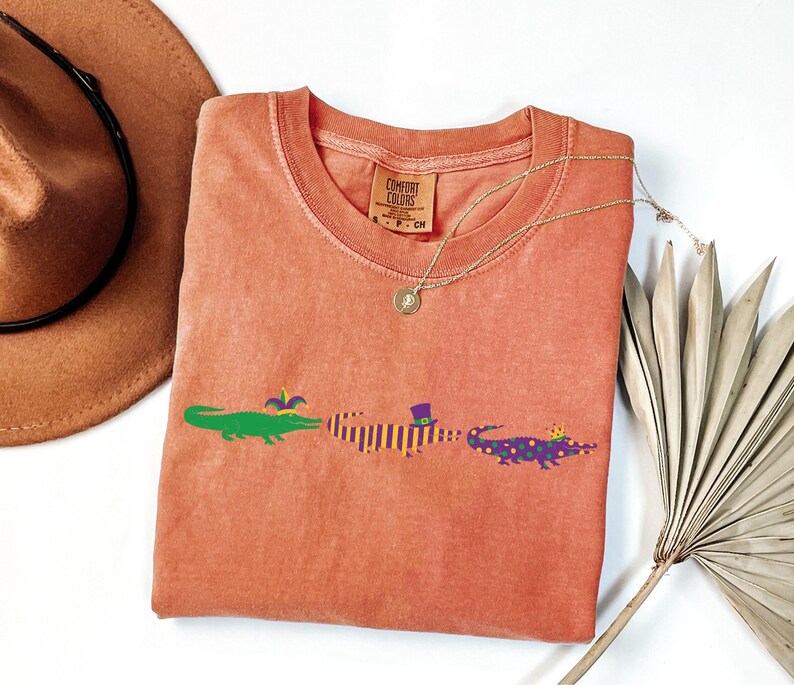 K&ouml;nnte beinhalten: Ein korallenfarbenes Comfort Colors T-Shirt mit drei Cartoon-Alligatoren in Gr&uuml;n, Lila und Lila mit goldenen Kronen. Die Alligatoren sind mit Accessoires zum Thema Mardi Gras verziert. Eine goldene Halskette und ein brauner Hut sind ebenfalls im Bild.