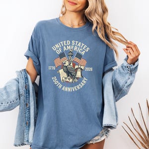 May include: A blue t-shirt with a graphic commemorating the 250th anniversary of the United States of America. The design includes an eagle, flags, and the text "United States of America 250th Anniversary 1776-2026."