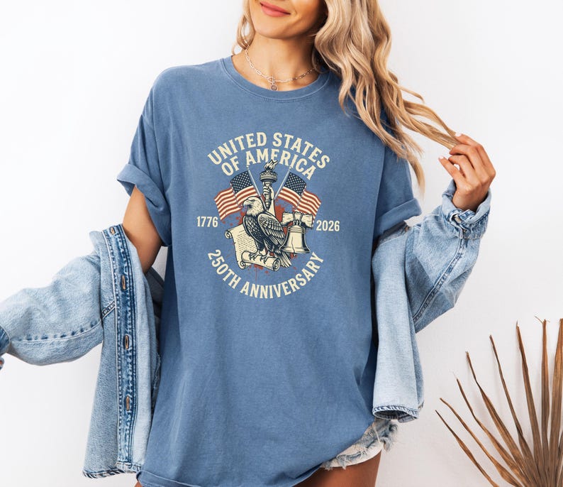 May include: A blue t-shirt with a graphic commemorating the 250th anniversary of the United States of America. The design includes an eagle, flags, and the text "United States of America 250th Anniversary 1776-2026."