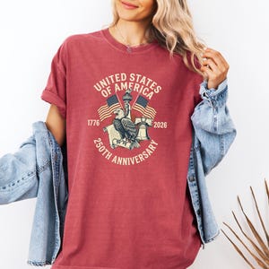 May include: A red t-shirt with a graphic commemorating the 250th anniversary of the United States of America. The design includes an eagle, flags, and the text "United States of America 250th Anniversary 1776-2026."
